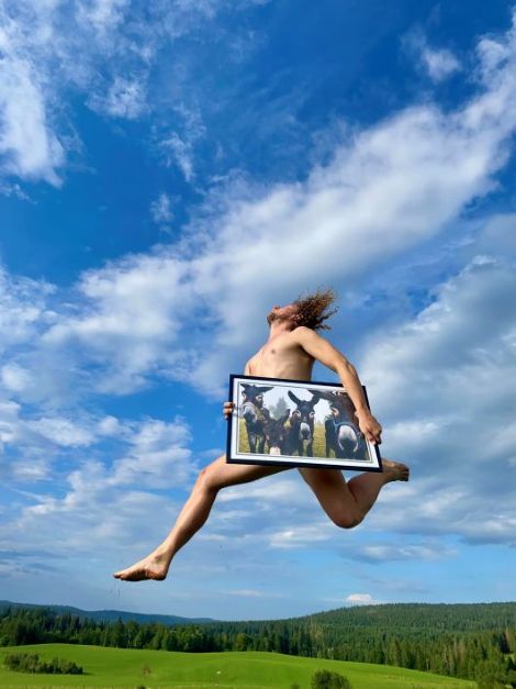 un ciel bleu avec quelques nuages et un champ vert. un homme saute nu, portant une photo